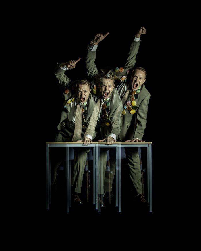 DETENTION: Three men leaning on a desk, looking angry and shouting, waving an arm in the air.