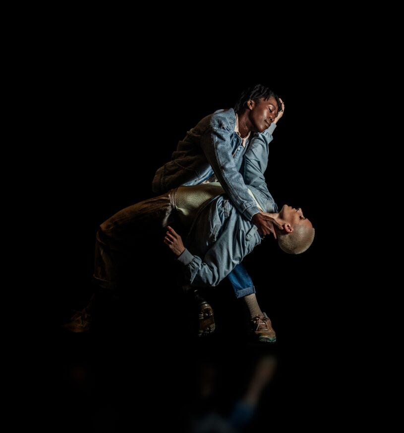 DETENTION: Two female dancers in blue denim jackets, one black the other with short white hair. One supporting the other in a deep bend, both looking tenderly.