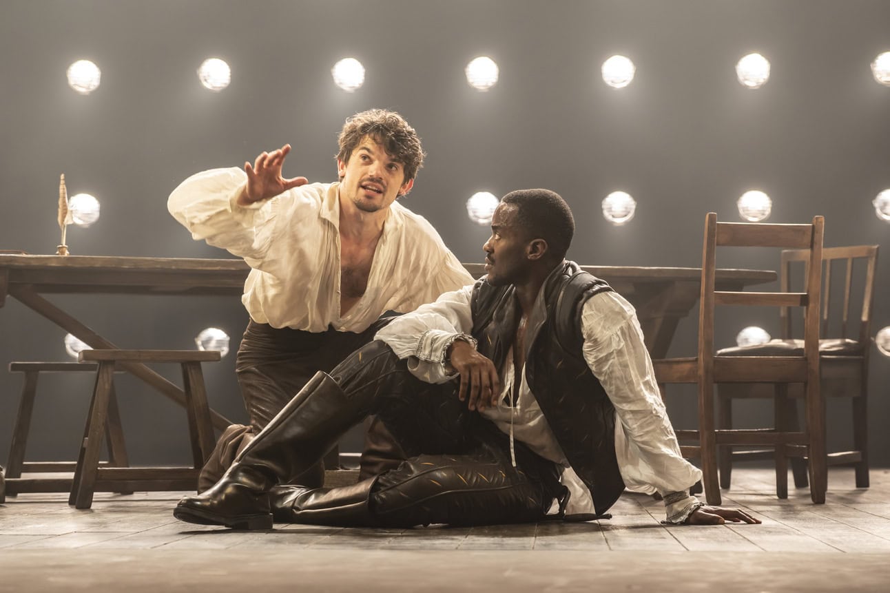 Edward Bluemel as William Shakespeare wearing a white linen shirt kneeling on the ground behind Ncuti Gatwa as Christopher Marlowe sitting on the ground wearing a jerkin and hose. 