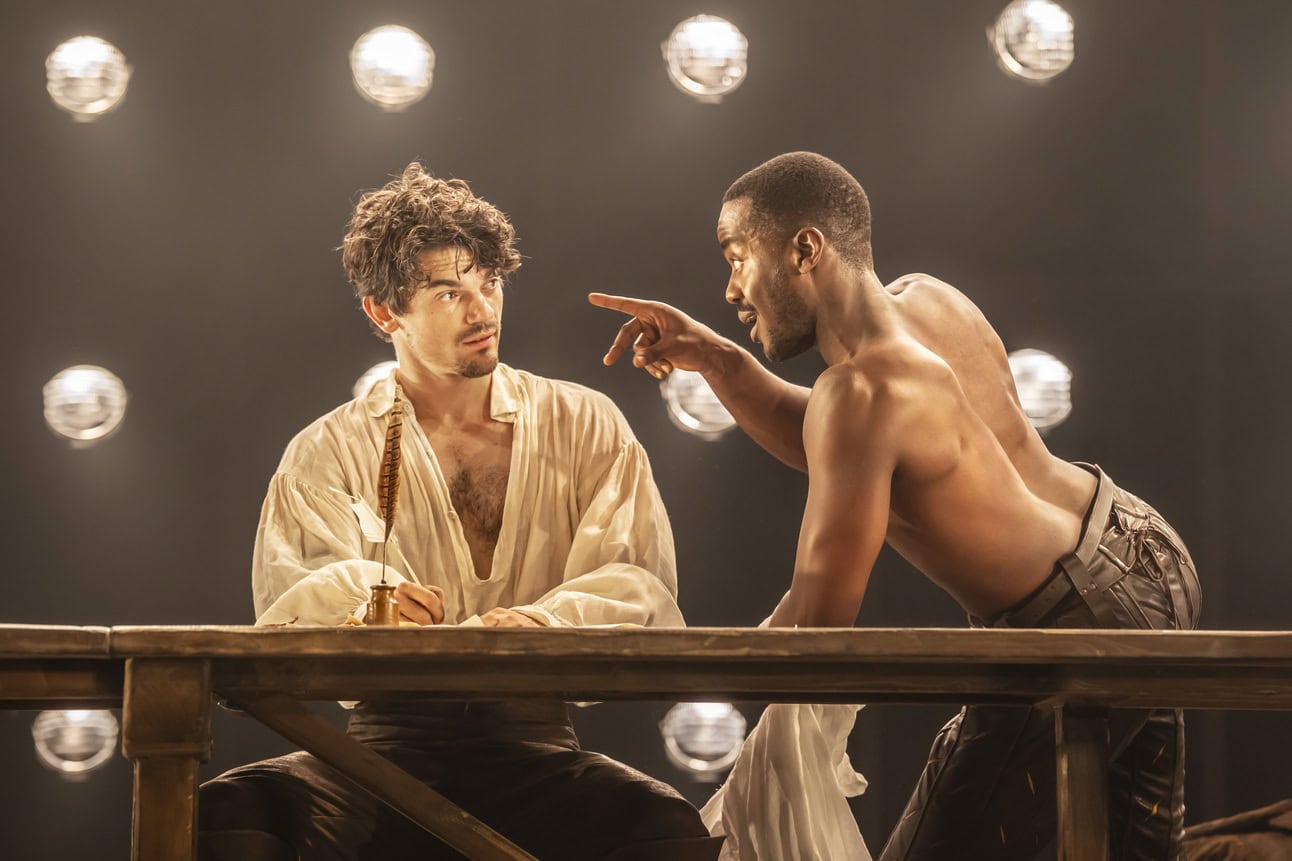 Edward Bluemel as William Shakespeare wearing a white linen shirt sitting at a wooden table and writing with a quill as Ncuti Gatwa as Christopher Marlowe is shirtless and leaning towards him and pointing a finger at him. 