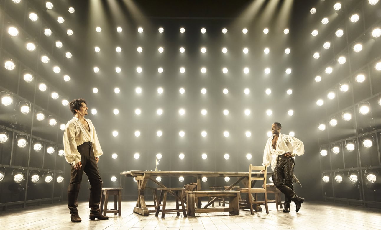 Edward Bluemel as William Shakespeare and Ncuti Gatwa as Christopher Marlowe standing across from each other on the far ends of a wooden table and looking at eachother.  