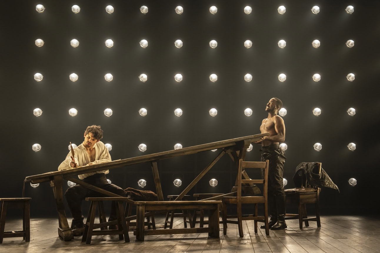 Edward Bluemel as William Shakespeare wearing a white linen shirt and writing at a wooden table with a quill as a shirtless Ncuti Gatwa lifts the table to an angle from the far side. 