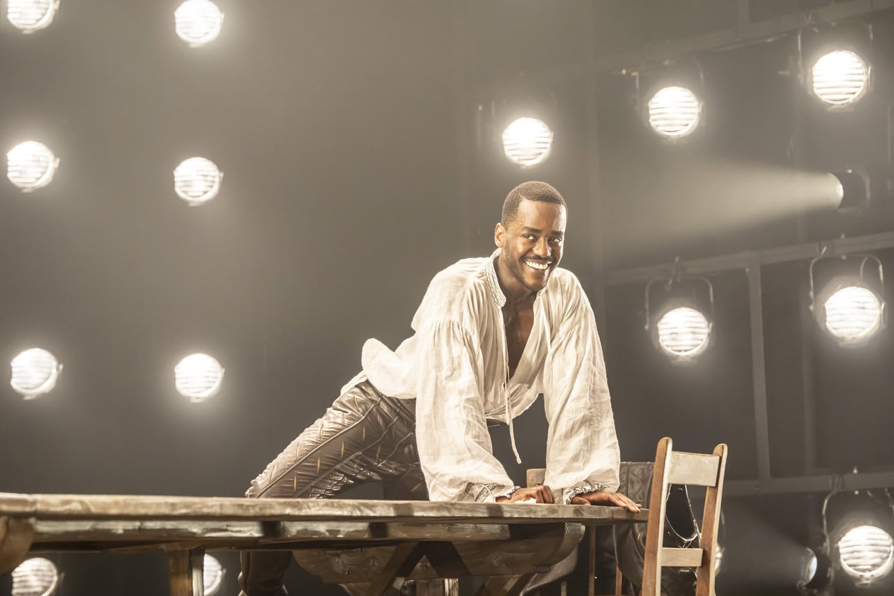 Ncuti Gatwa as Christopher Marlowe wearing a white linen shirt and leather hose climbing on a wooden table and smiling. 