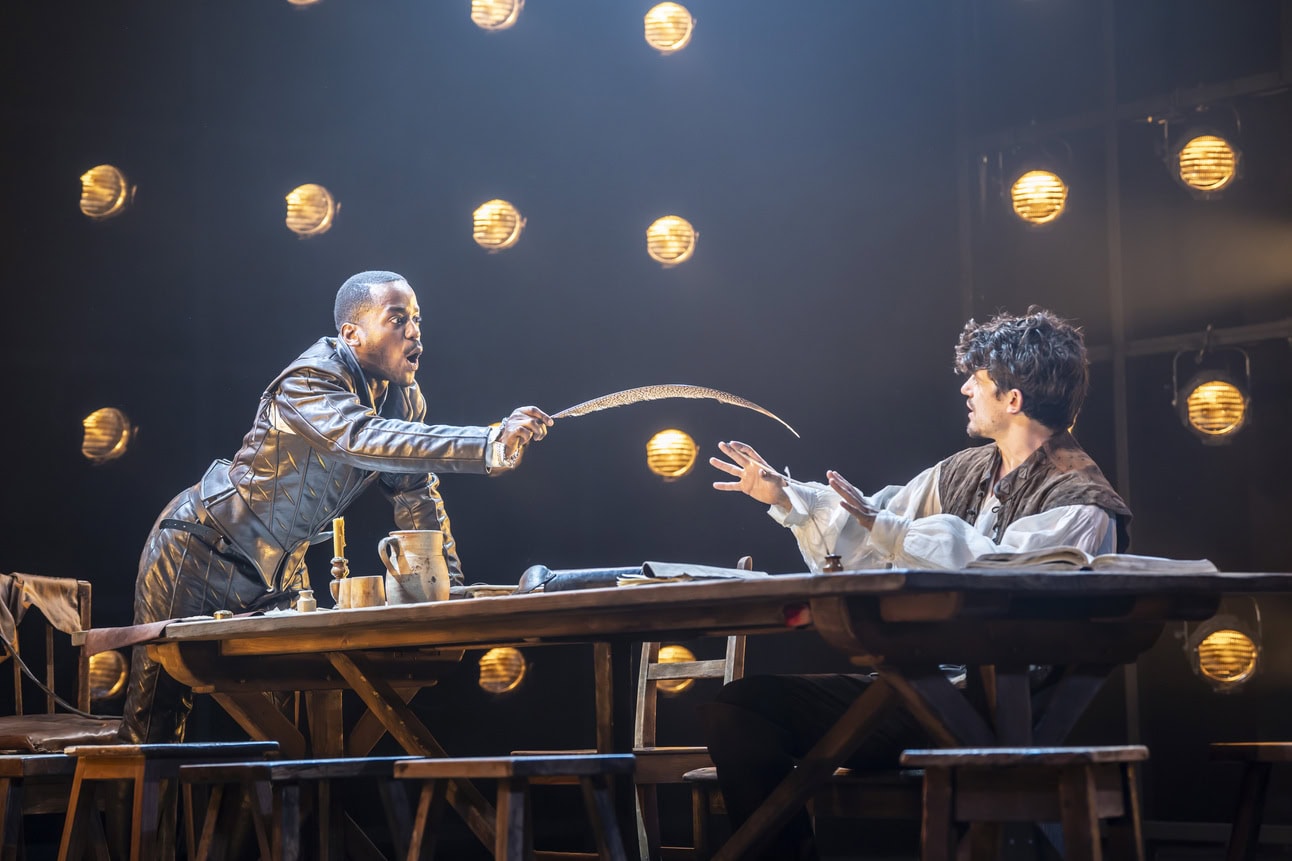 Ncuti Gatwa as Christopher Marlowe wearing a leather doublet while pointing a quill and leaning towards Edward Bluemel as William Shakespeare sitting at the table and looking at him. 