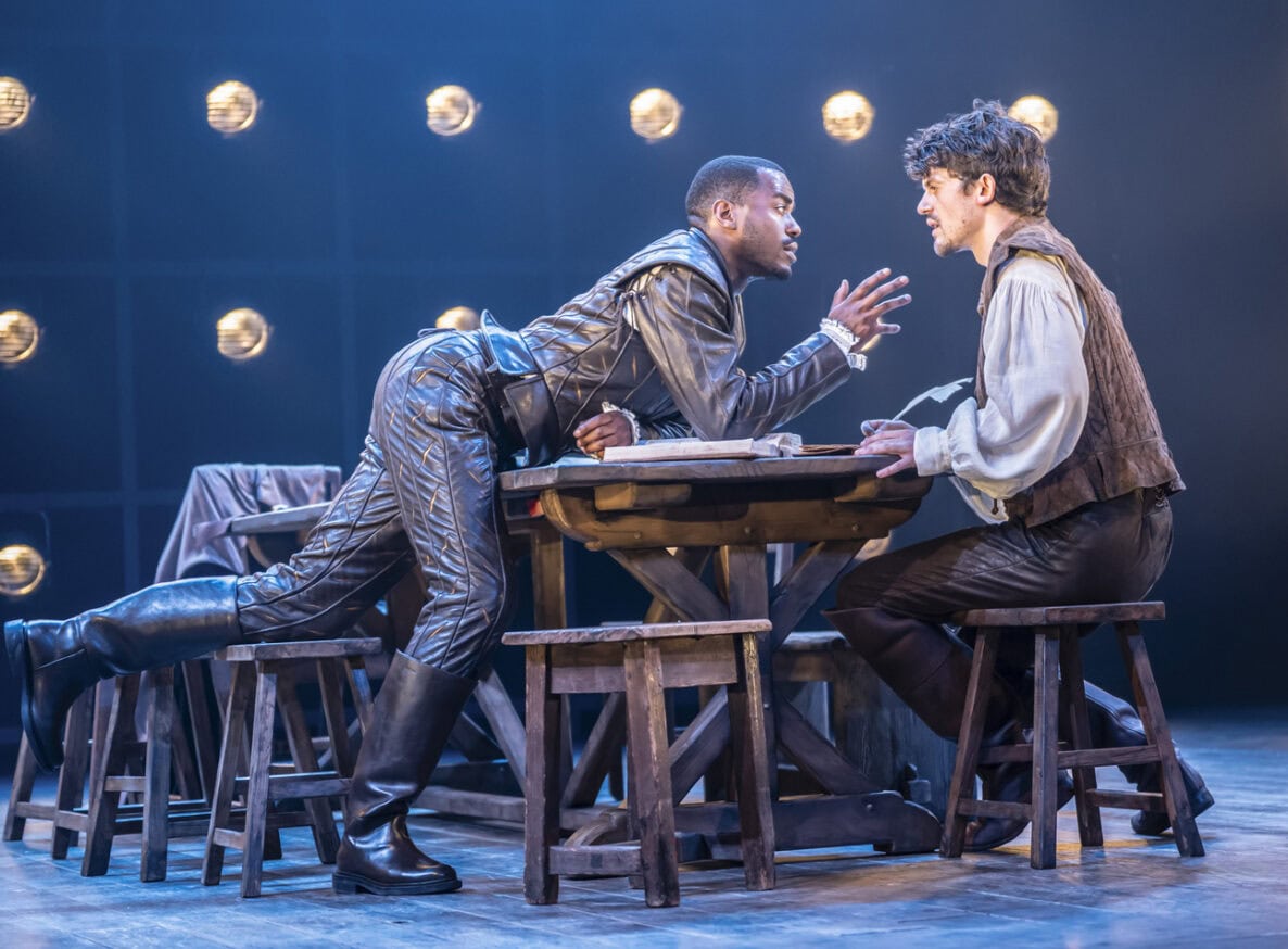 Ncuti Gatwa as Christopher Marlowe wearing leather doublet and hose and leaning over a wooden table towards Edward Bluemel as William Shakespeare wearing a white linen shirt and a brown jerkin sitting at the table and writing. 