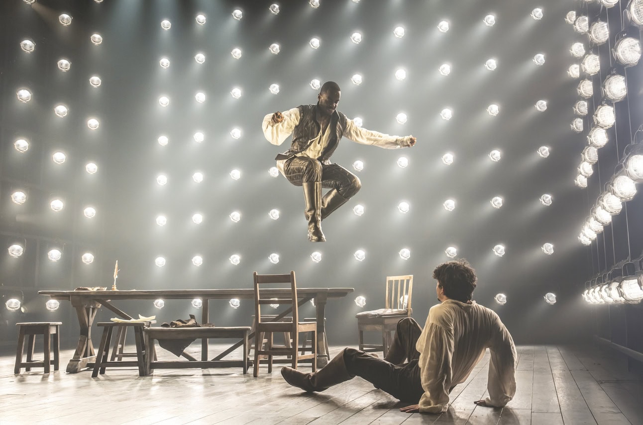 Ncuti Gatwa as Christopher Marlowe wearing a jerkin, white linen shirt and hose mid-air jumping off a table towards Edward Bluemel as William Shakespeare on the ground looking up at him wearing a white linen shirt. 
