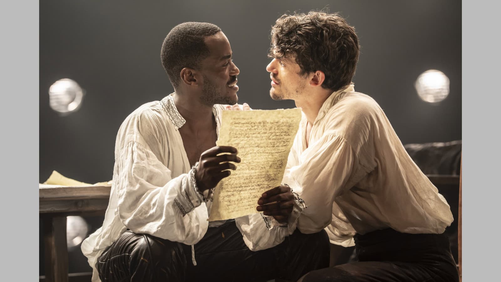 Ncuti Gatwa as Christopher Marlowe and Edward Bluemel as William Shakespeare wearing white linen shirts and sitting together and looking at each other while holding a piece of paper.