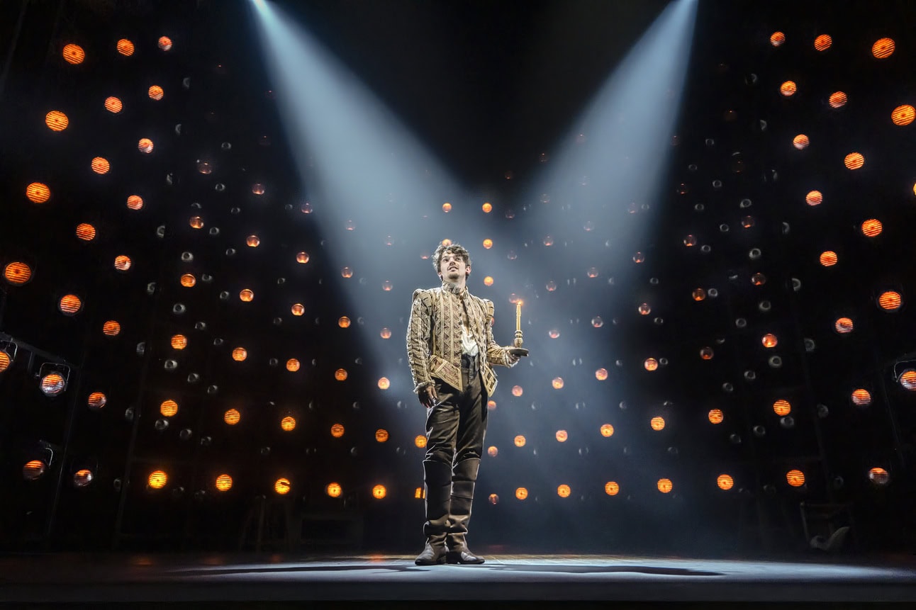 Edward Bluemel as William Shakespeare wearing doublet and hose, holding a candle and standing in the spotlight with walls or orange lights behind him. 