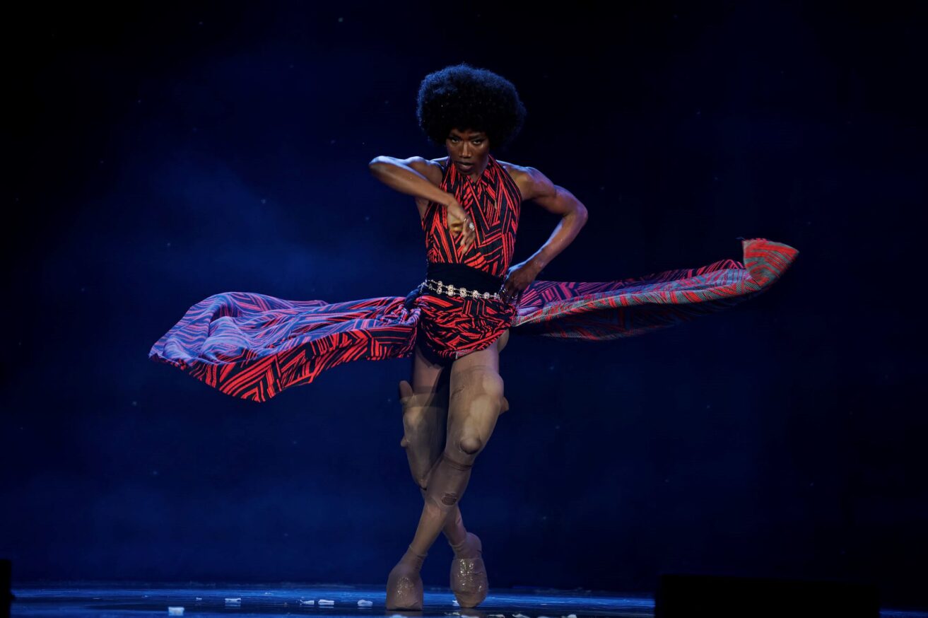 A performer wearing a red and black dress, swishing their skirt dramatically and facing the camera